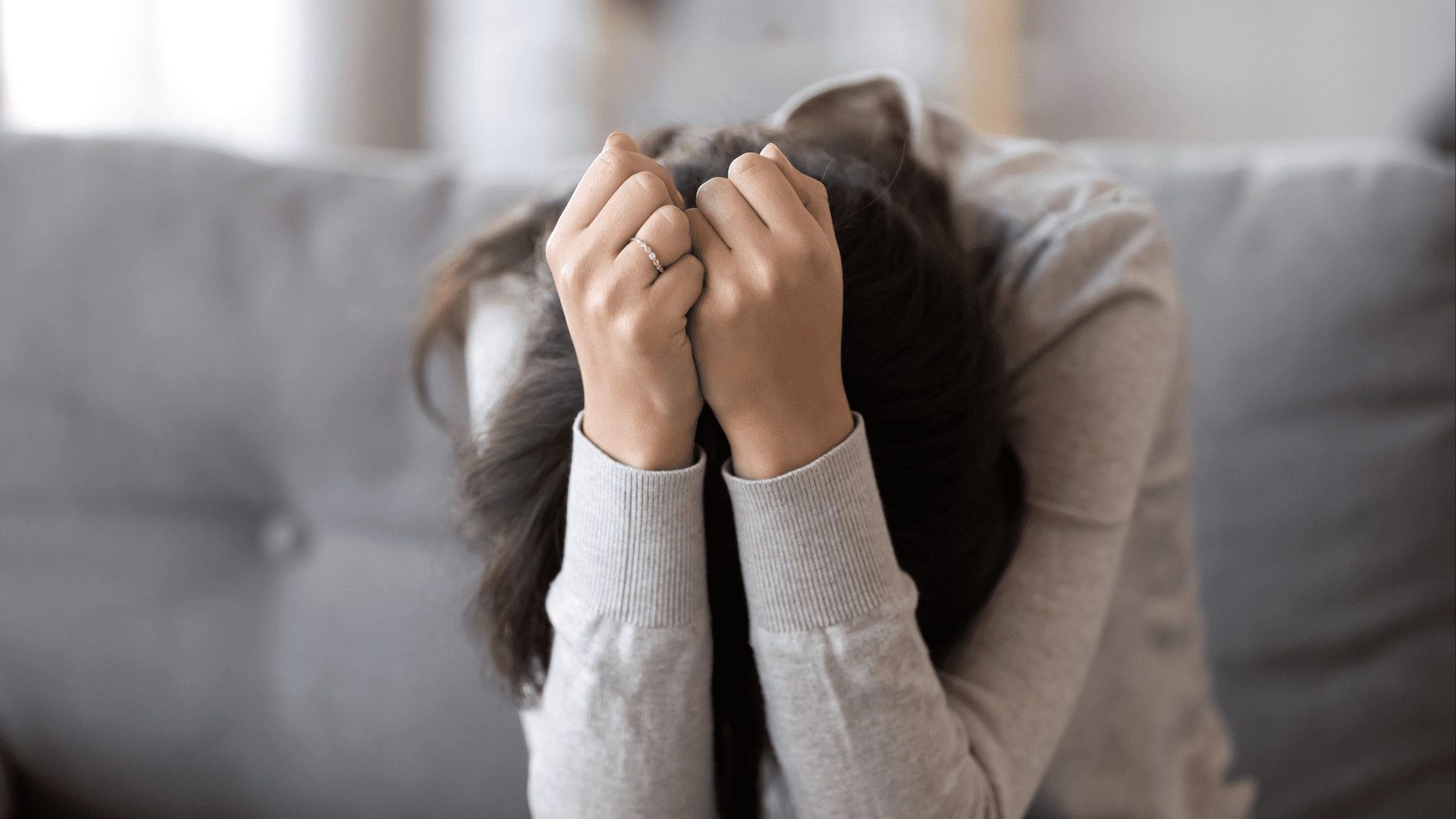 a woman sitting on a couch wuth her head in. her hands who is depressed and wondering what causes depression