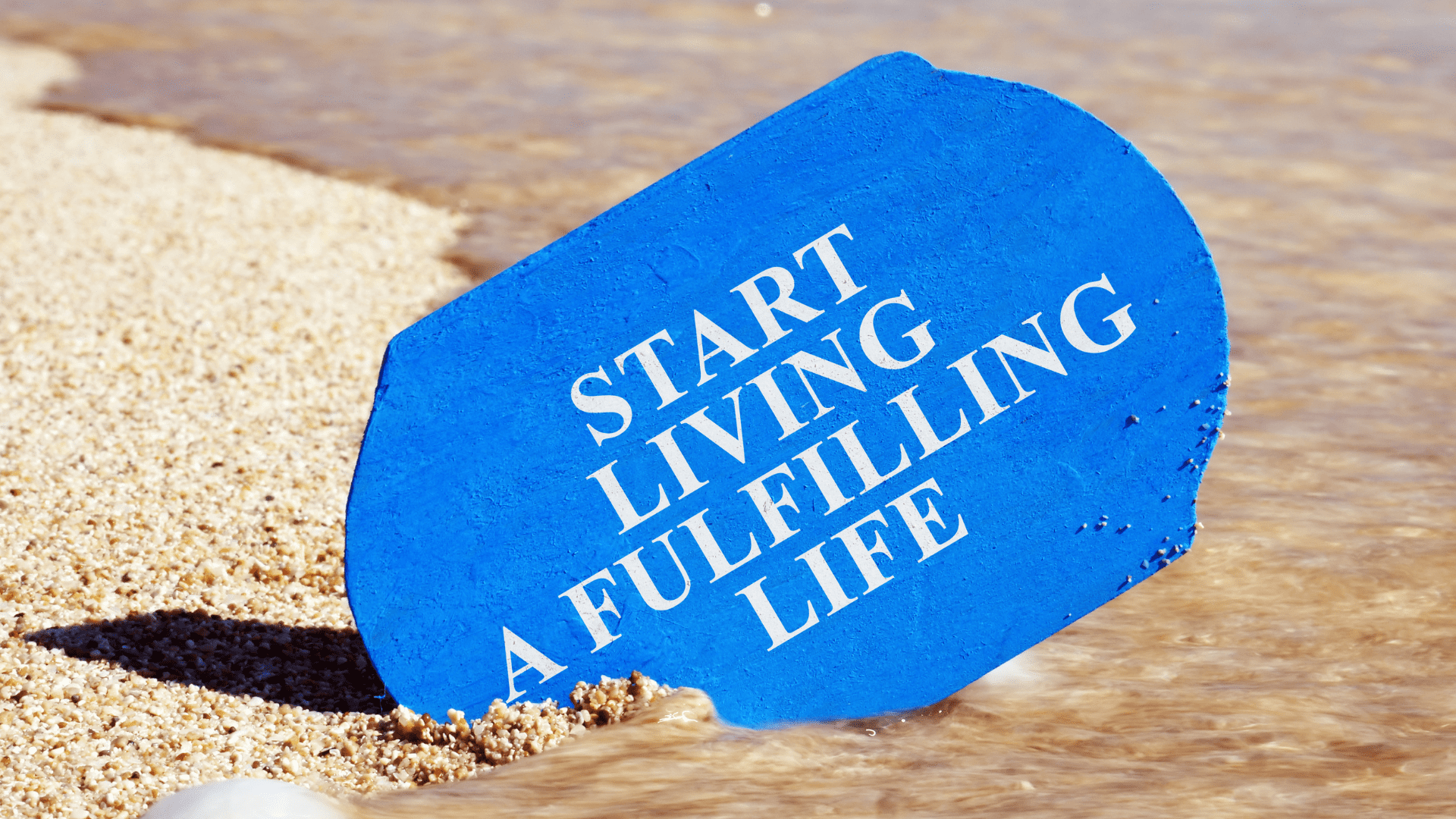 Blue sign in the sand of a beautiful beach with the text 'Start Living a Fulfilling Life' representing the importance of positive habits that lead to a fulfilled life, life coaching, and feeling fulfilled in life.