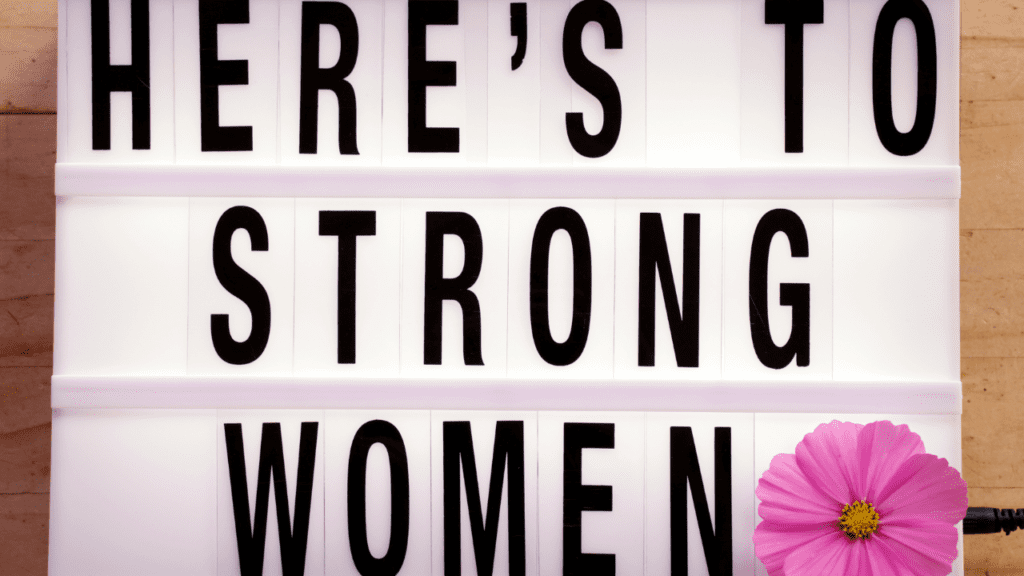 Woman holding a sign saying "Here's to strong women" symbolizing the balance of strength and empowerment in women.