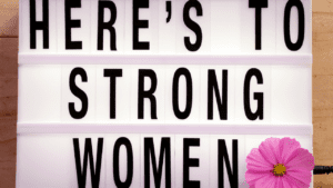 Woman holding a sign saying "Here's to strong women" symbolizing the balance of strength and empowerment in women.