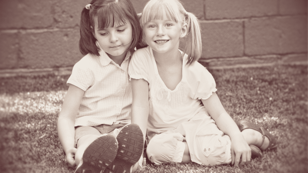 Two young girls sitting together, one smiling and the other looking down from experiencing childhood trauma.