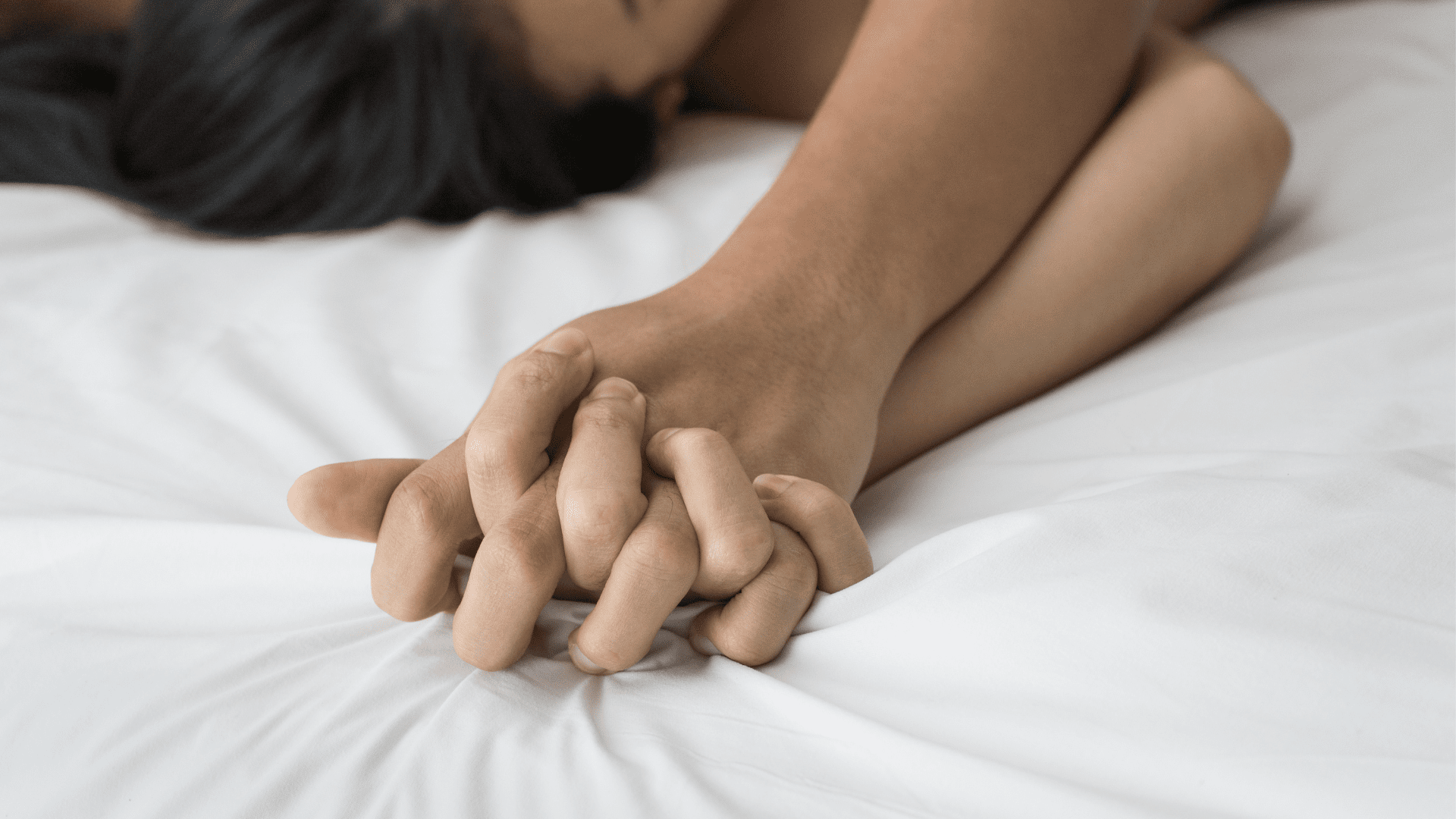 Close-up of intertwined hands on a bed, symbolizing intimacy.