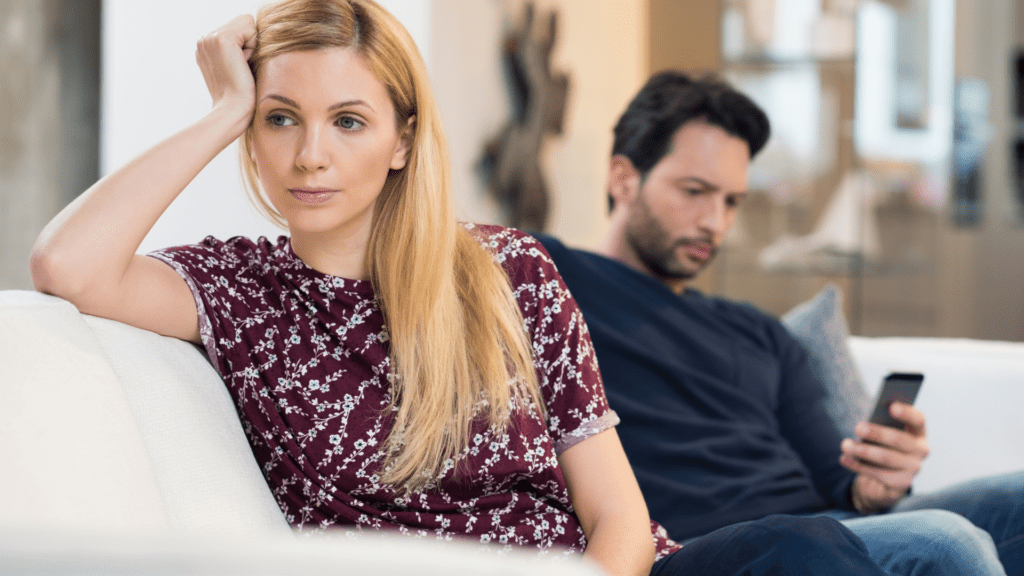 Woman looking frustrated while partner focuses on phone when she needs him to be emotionally available.

A woman in a burgundy floral dress sits on a white couch looking frustrated and disconnected while her partner in the background is absorbed in his phone, illustrating the impact of digital distraction on emotional availability.