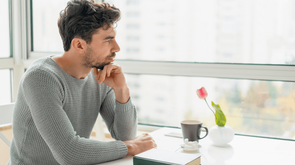 Man sitting at a table, looking out the window with a thoughtful expression, symbolizing introspection and emotional struggle.

