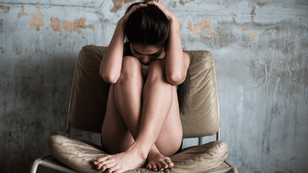 Woman sitting curled up on a chair, holding her head in distress against a worn-out wall.

