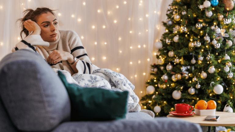 Woman sitting on a couch next to a Christmas tree, looking sad and lost in thought.