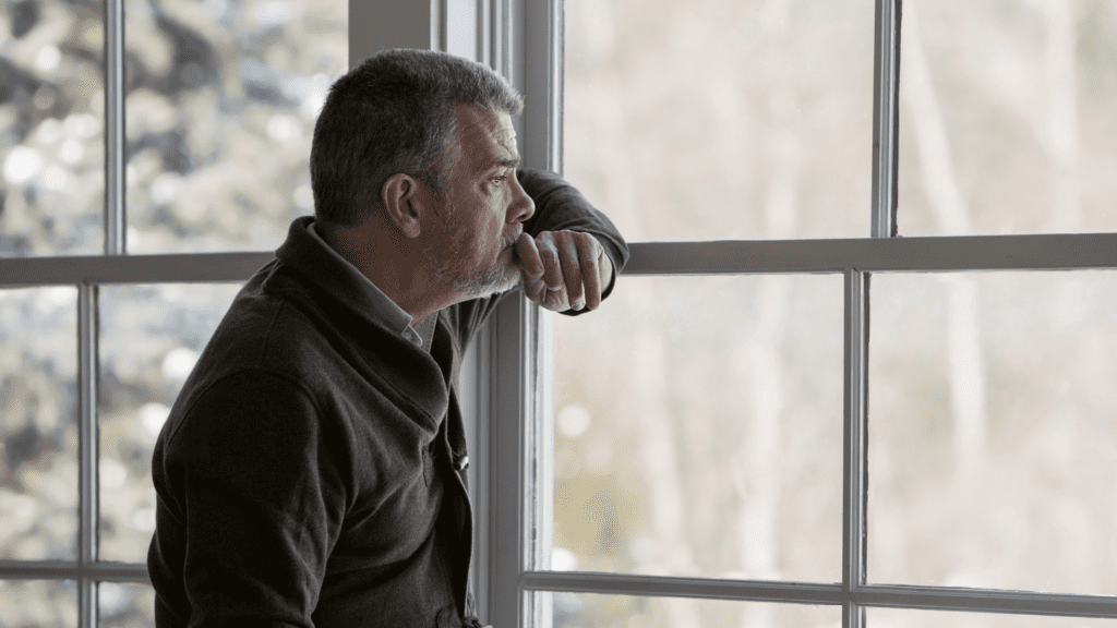  Elderly man looking out a window during the winter, appearing thoughtful and melancholic.

 