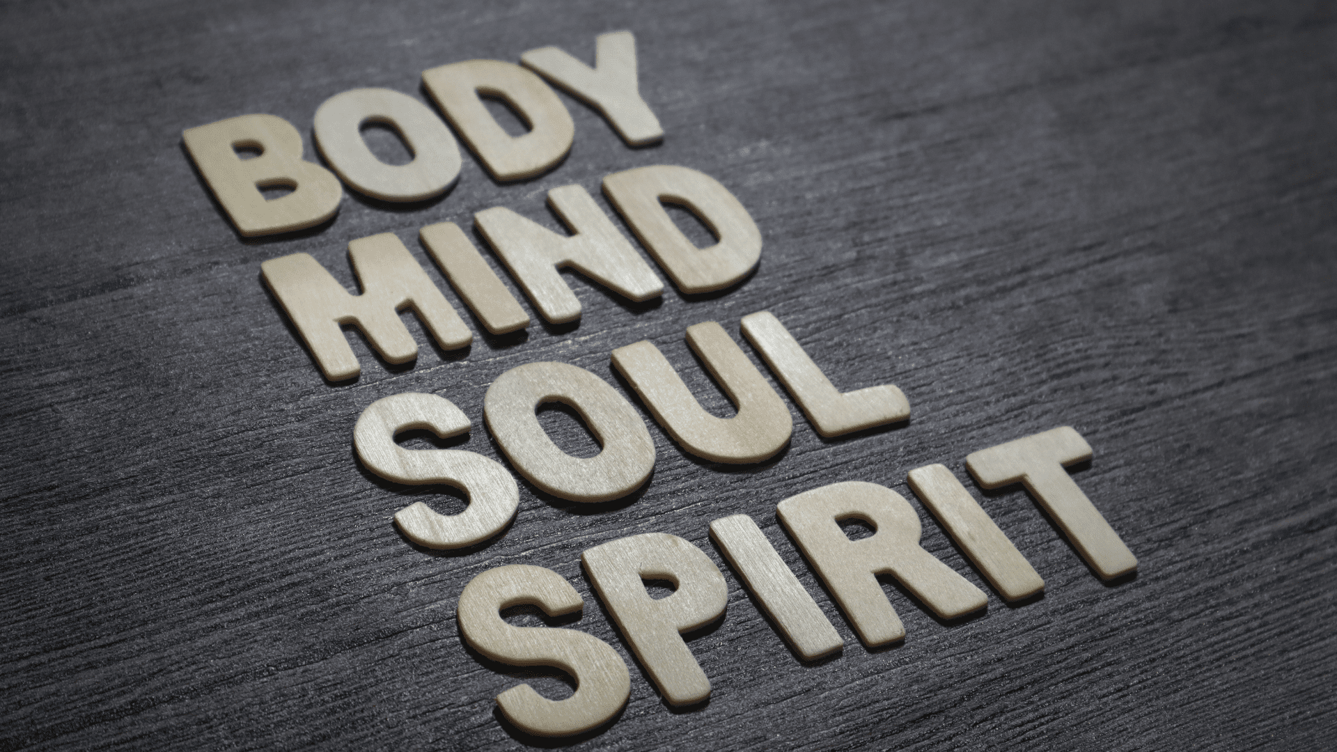 Wooden letters spelling "Body, Mind, Soul, Spirit" on a dark wooden surface. Wooden letters arranged on a dark wooden surface spell out the words "Body, Mind, Soul, Spirit," emphasizing the foundation of holistic psychology in addressing all aspects of human well-being.