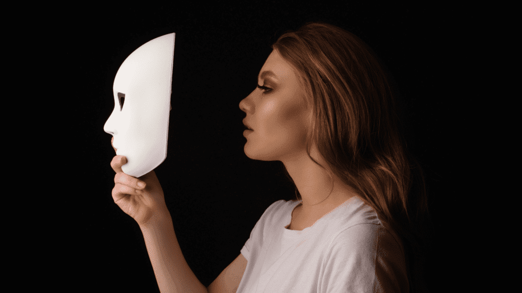 Woman in profile holding white mask against dark background, symbolizing hidden identity

