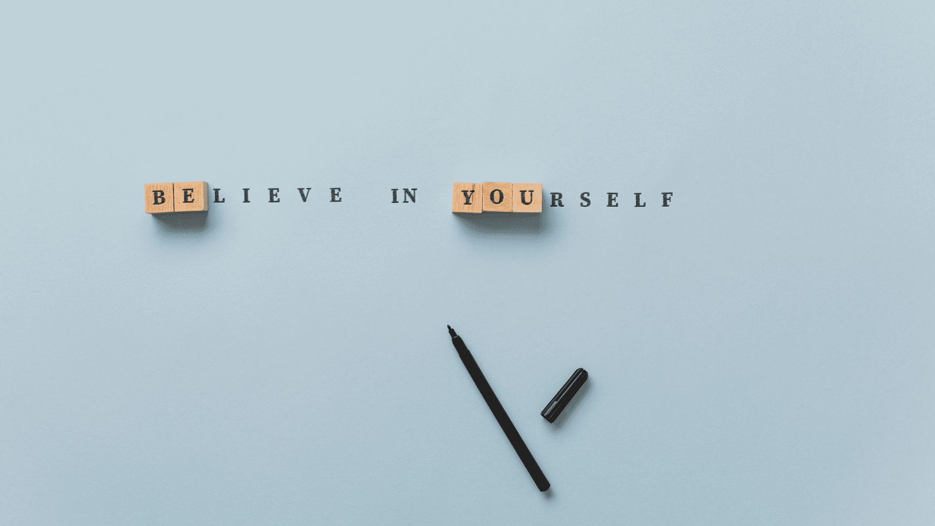 Wooden letter blocks spelling "Believe in Yourself" on a minimalist blue background with a black pen beside them.