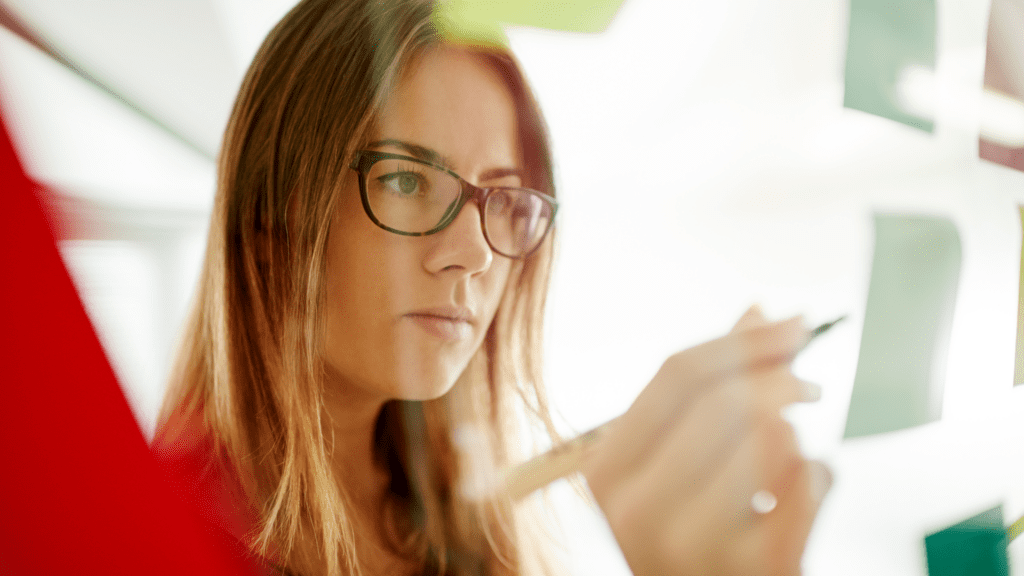 A woman with glasses intently writing on a transparent board, embodying a strategic visionary mindset.

