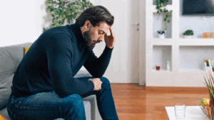 A man sitting on a couch with his head in his hand, showing signs of stress and fatigue linked to dopamine depletion.