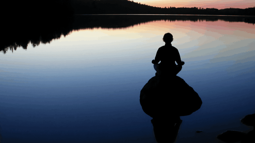 Person meditating by a calm lake at sunset, symbolizing mindfulness and neuroplasticity through peaceful awareness.