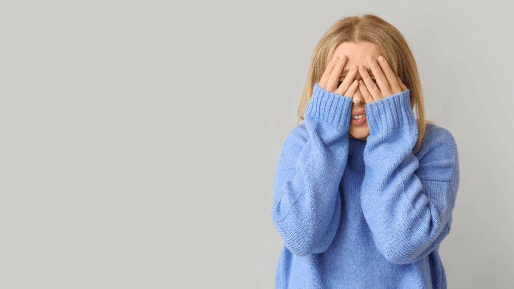 Woman in blue sweater covering her face, expressing insecurity about feeling like a loser.